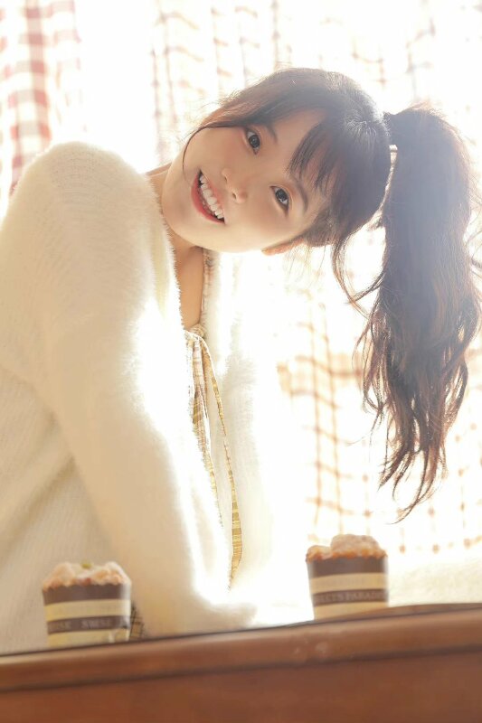 Tạo dáng đứng nghiêng người hơi cúi với tóc đuôi gà, mặt quay nghiêng, 2 tay chống lên bàn mắt nhìn ống kính miệng cười tươi siêu cute