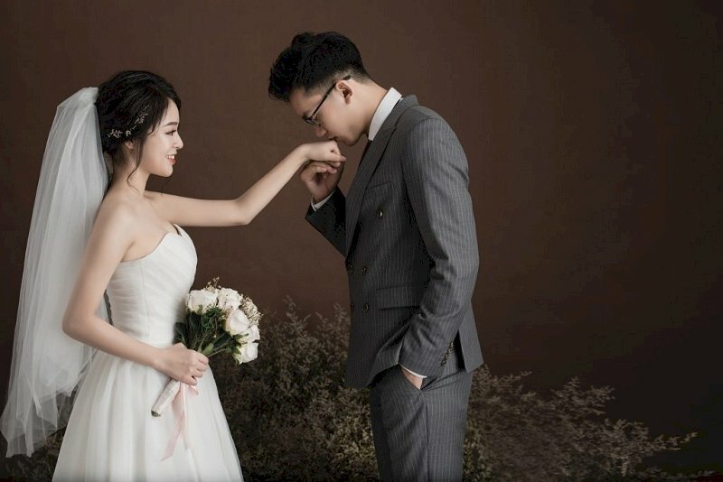 Cách tạo dáng chụp ảnh cưới trong studio đơn giản ngọt ngào bằng cách chú rể cầm  và hôn lên tay cô dâu, 1 tay thì bỏ vào túi phần, cô dâu 1 tay cầm bó hoa để ngang bụng.