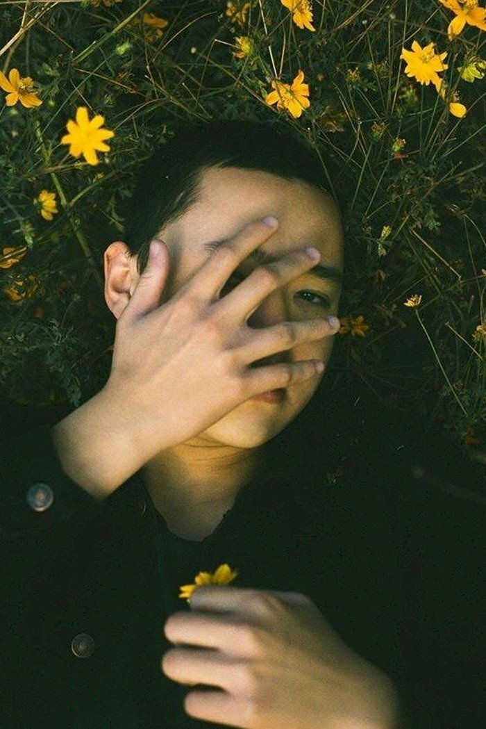 Cách tạo dáng chụp ảnh giấu mặt nam với background hoa cỏ, mẫu nam nằm xuống, 1 tay cầm bông hoa còn tay còn lại đưa lên che mặt. 