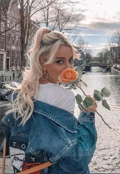 Tạo dáng cô gái trong bộ quần áo mang phong cách năng động, 1 tay cầm nhánh hoa hồng che nửa mặt dưới, bức ảnh được chụp lấy nửa người góc chụp từ sau lưng