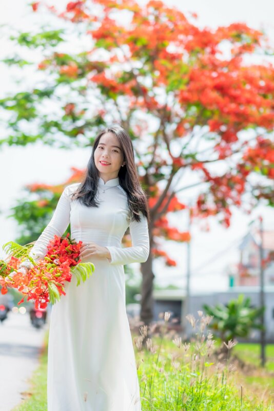 Tạo dáng đứng xinh tươi trong chiếc áo dài trắng học sinh với 1 tay ôm nón lá chứa hoa phượng đỏ, một tay nắm lấy vành nón, người hơi nghiêng miệng cười tươi mắt nhìn ống kính