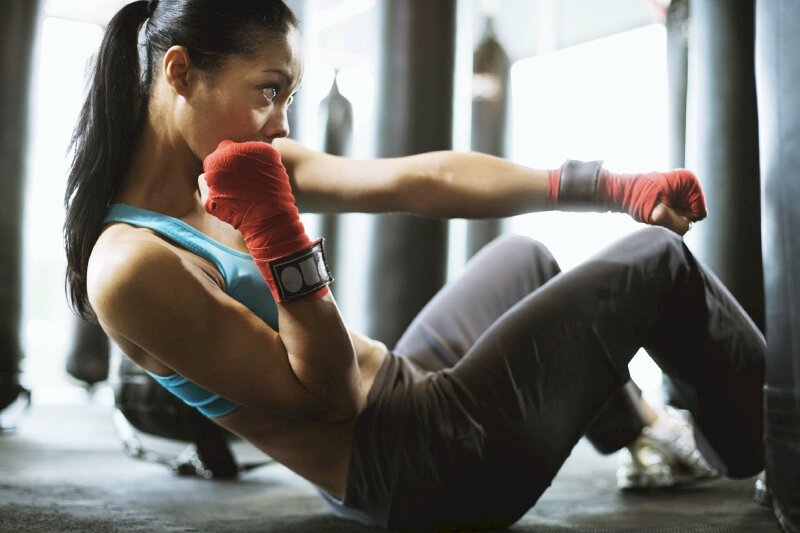 Tạo dáng gymer nữ đang đánh boxing trong tư thế ngồi gập bụng, chụp góc nghiêng, đầu hơi cúi, mắt nhìn theo nắm đấm thể hiện sự máu lửa