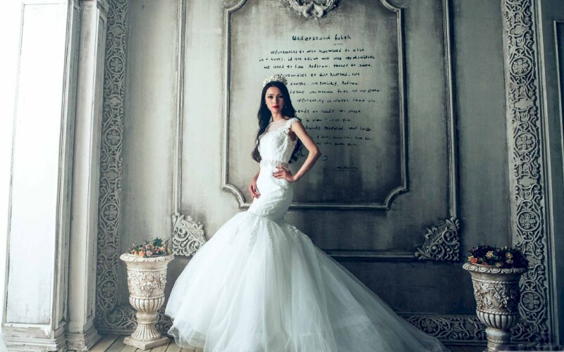 Tạo dáng váy cưới chụp chính diện, mắt nhìn ống kính, một tay chống hông, tay còn lại chạm nhẹ vào đùi