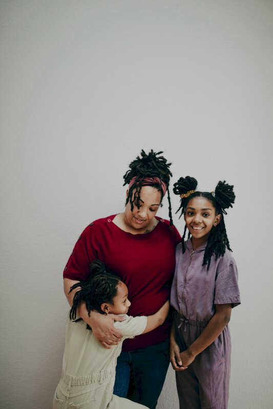 Tạo dáng gia đình 3 mẹ con đầm ấm với một bé ôm mẹ, một bé đứng nghiêng người hướng vào mẹ, miệng cười tươi nhìn ống kính