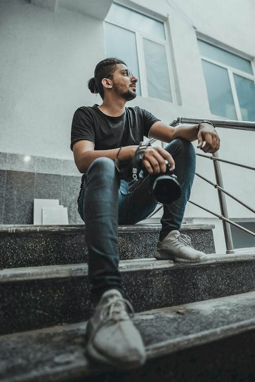 Cách tạo dáng chụp ảnh cho nam khi ngồi cầu thang ngầu chất với góc chụp từ dưới lên, ngồi đặt 1 chân trước hướng về phía ống kính và 1 chân sau, 1 tay đặt lên đầu gối, 1 tay cầm máy ảnh, đầu ngước lên nhìn phía trên