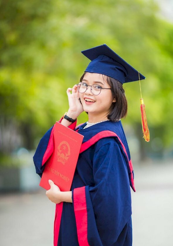 Kiểu chụp cute cho các bạn nữ trong bộ áo cử nhân với góc chụp nghiêng, dáng đứng thẳng, một tay vén tóc, một tay cầm bằng, miệng cười tươi