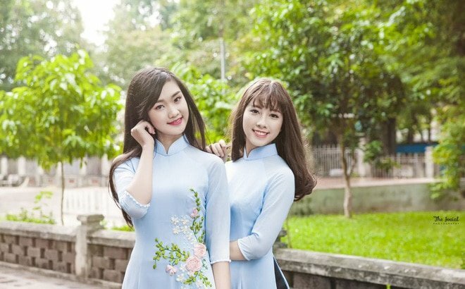 Cách tạo dáng chụp kỷ yếu lưu giữ thanh xuân dành cho 2 bạn nữ sinh viên trong bộ áo dài xanh nhạt với cách chụp nửa người, một bạn đưa tay vén tóc, một bạn đứng nép về phía sau tay chạm nhẹ lên vai bạn, miệng cười tươi, mắt nhìn ống kính