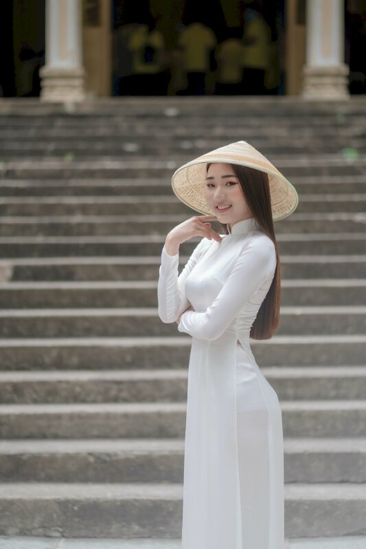 Cách tạo dáng đầy duyên dáng và dễ thương với góc chụp nghiêng người, 1 tay đặt trước bụng, 1 tay chạm nhẹ vào má, mắt nhìn ống kính, miệng cười tươi