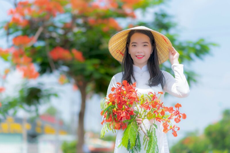Cách tạo dáng đứng đẹp tự nhiên với một tay cầm bó hoa phượng, một tay chạm vành nón là, mắt nhìn thẳng ống kinh, miệng cười tươi