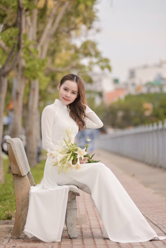Dáng ngồi ghế đá dịu dàng với một tay ôm bó hoa ly, một tay vén tóc xõa một bên, miệng cười nhẹ, mắt nhìn ống kinh