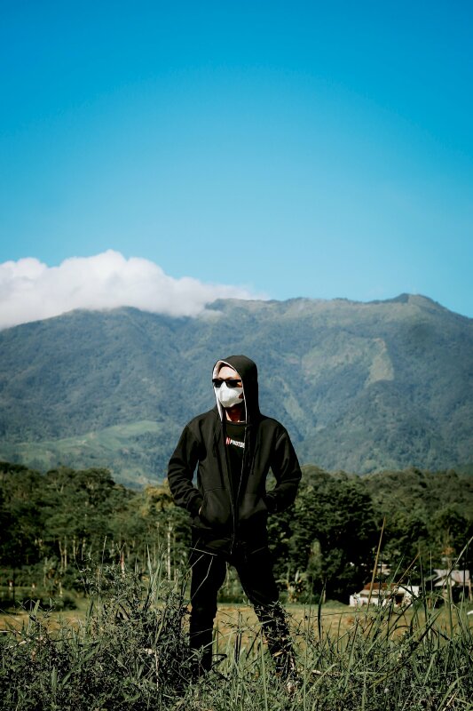 Kiểu chụp ảnh chill với thiên nhiên với bộ quần áo đen, mang khẩu trang và kính đen, mặt nhìn nghiêng, 2 tay đặt túi áo, 2 chân đứng rộng bằng vai