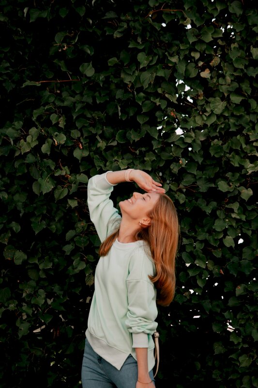 Kiểu chụp ảnh nửa người với góc chụp nghiêng trên nền background lá xanh biếc cùng dáng người đầu ngẩng lên cao, mắt nhắm, miệng cười tươi