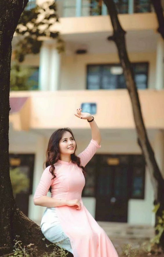 Kiểu dáng ngồi tự nhiên rất đáng yêu trên góc cây to của sân trường, tay đặt trước bụng, tay dơ cao như che nắng, mặt ngước lên, miệng cười tươi