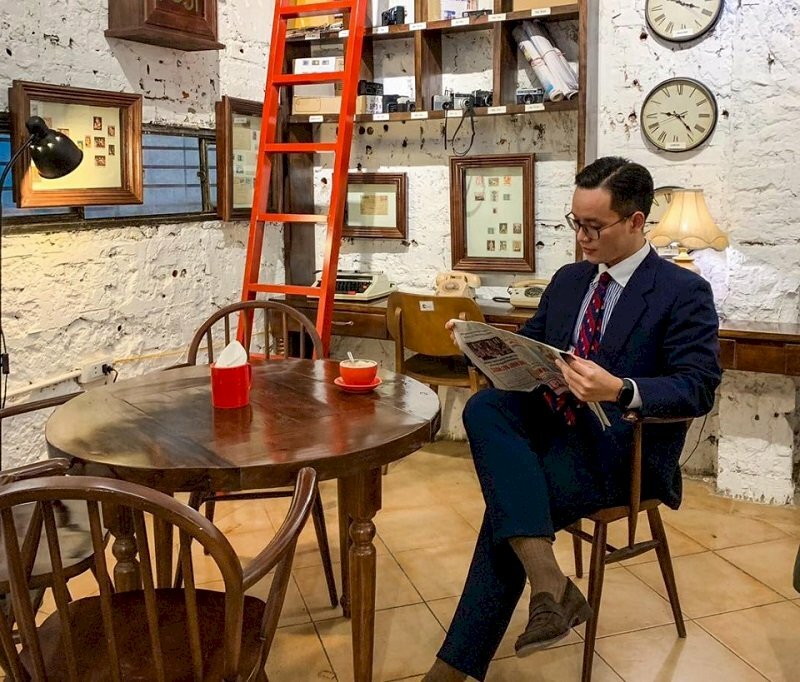 Kiểu tạo dáng đàn ông trong bộ áo vest sang trọng đang ngồi bắt chéo chân xem báo cùng trên bàn là tách cafe capuchino