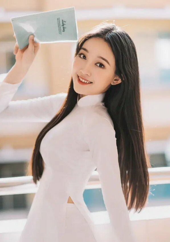 Kiểu tạo dáng xinh tươi với góc chụp nghiêng tay để phía sau tay còn lại cầm sách che trên trán, miệng cười tươi, mắt nhìn ống kính