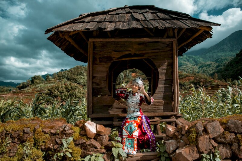 Tạo dáng đẹp trong tư thế ngồi tự nhiên, 2 tay chạm mũ, đầu hơi nghiêng, miệng cười tươi, mắt nhìn ống kính