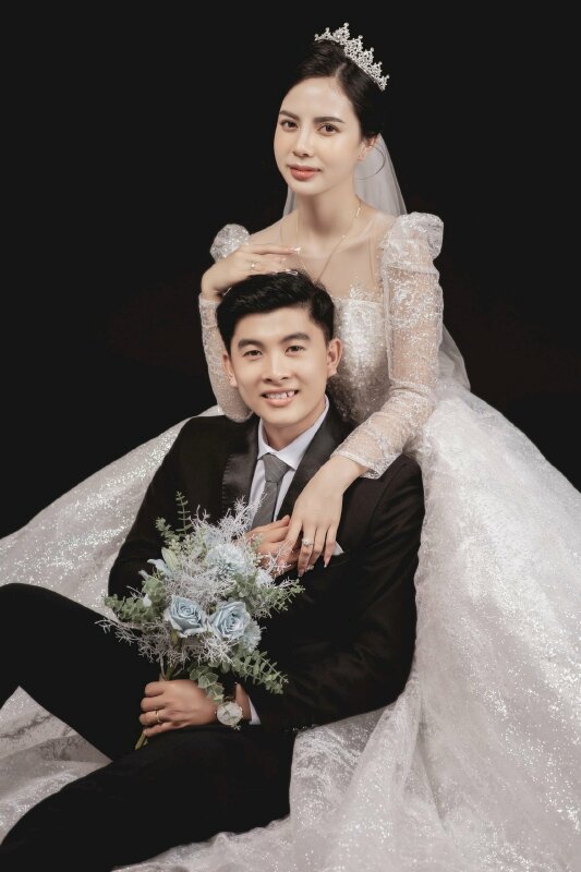 Cách tạo dáng chụp ảnh cưới trong studio tình tứ với cô dâu ngồi trên ghế chú rể ngồi dứa nền dựa vào cô dâu, 1 tay cô dâu đặt qua vai chú rể và chú rể đưa tay cầm lấy tay cô dâu.