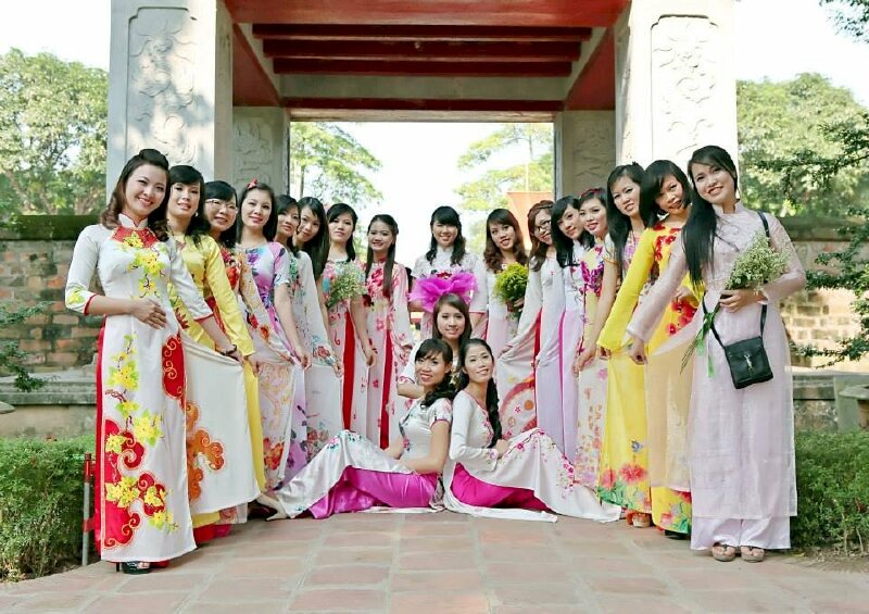 Tạo dáng chụp ảnh kỷ yếu sinh viên cho nhóm nhiều bạn hữ trong bộ áo dài hoa tuyệt đẹp, chính giữa là 2 bạn nữ ngồi tựa lưng vào nhau, cùng một bạn ngồi phía sau và một bạn cầm bó hoa, các bạn còn lại người hơi nghiêng, tay cầm vạt sau áo dài, miệng cười tươi