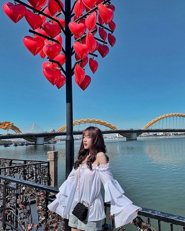 Cách tạo dáng chụp ảnh ở cầu Tình Yêu đẹp lấy được view cầu rồng, với góc này bạn hãy đứng dưới cây cột có lòng đèn trái tim, và đứng xoay lưng người về phía cầu rồng, tựa lưng vào thành cầu, 2 tay dang nhẹ hai bên cầm vào thành cầu, mắt nhắm nhẹ, là sẽ có được bức ảnh tuyệt đẹp ở cầu Tình Yêu. 