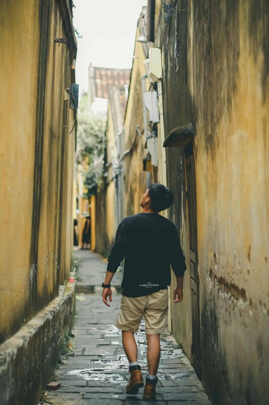 Tạo dáng chụp từ sau lưng với dáng bước đi tự nhiên giữa 2 bên là vách tường cổ kính, riêu phong, 2 tay xuôi thẳng tự nhiên, đầu hơi ngước nhìn