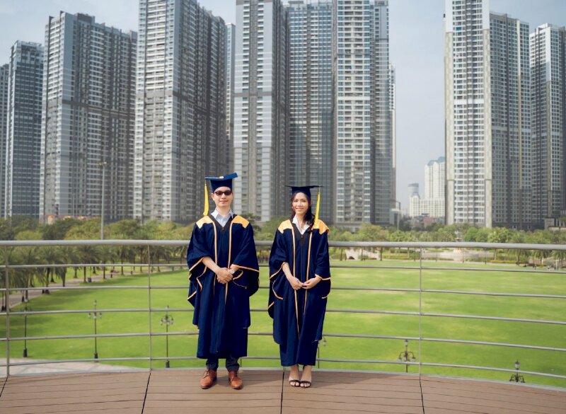 Tạo dáng siêu chất với đôi bạn trẻ trong bộ áo cử nhân trong tư thế đứngtrang nghiêm tay nắm chạt để phía trước, người đứng thẳng tự nhiên