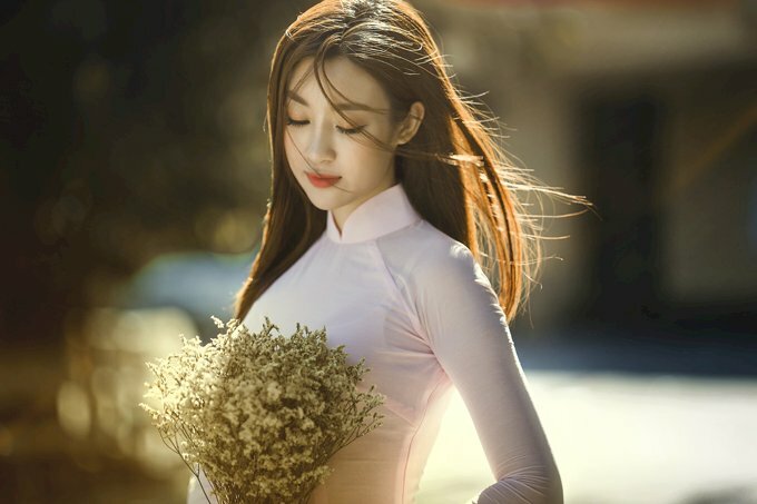 Tạo dáng nữ đẹp, đáng yêu trong chiếc áo dài trắng hồn nhiên, 2 tay cầm bó hoa dại khô, đầu hơi nghiêng, mắt nhìn hoa