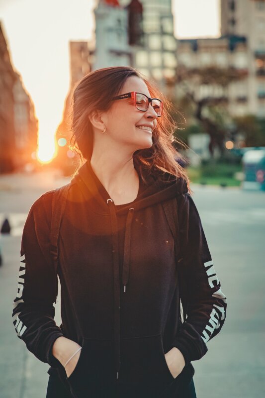 Tạo dáng thoải mái trong chiếc áo khoác màu đen 2 tay đặt vào túi áo, mặt hướng nghiêng cười tươi dưới bầu trời hoàng hôn từ phía sau lưng