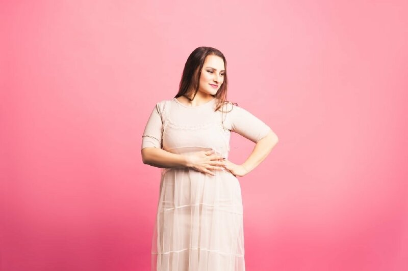 Tạo dáng chụp ảnh che bụng cho các cô gái mủm mỉm với dáng đứng thẳng tự nhiên, một tay chống hông, một tay che bụng mặt nhìn ngang, đầu hơi cứi, miệng cười nhẹ kết hợp cùng chiếc đầm suông dịu dàng, xinh xắn