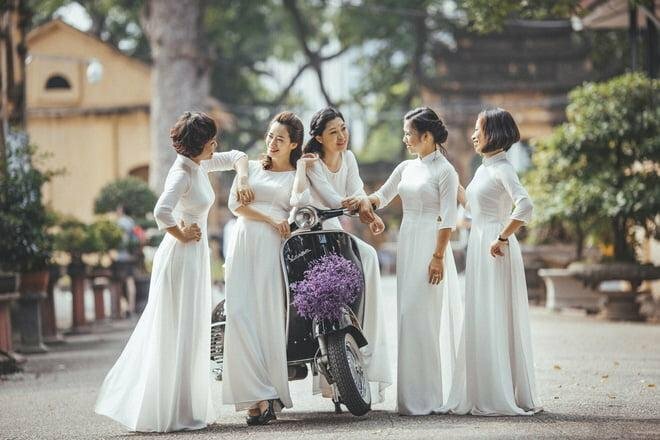 Cách tạo dáng chụp ảnh áo dài tập thể kiểu tự nhiên dành cho nhóm bạn 5 người, cùng nhau mặc áo dài trắng, với đạo cụ là chiếc xe vespa cổ màu đen có gắn chùm hoa tím, 1 người ngồi trên xe, 4 người đứng dưới, cả nhóm cùng đang trò chuyện vui vẻ.