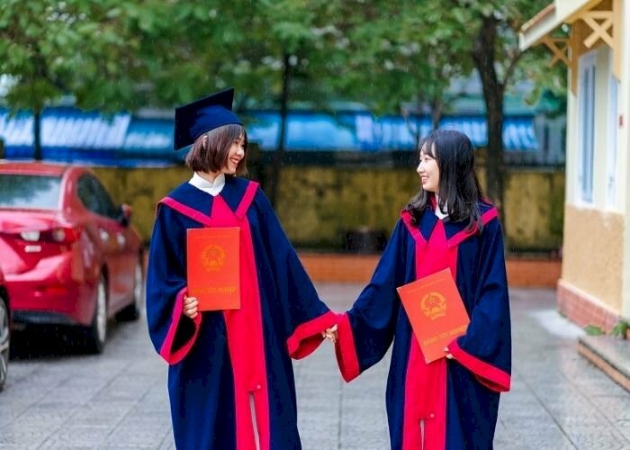 Cách tạo dáng chụp ảnh áo cử nhân kỷ yếu đôi nữ đang cầm tay nhau, hai gương mặt cùng quay vào nhìn nhau cười, tay cầm tấm bằng tốt nghiệp.