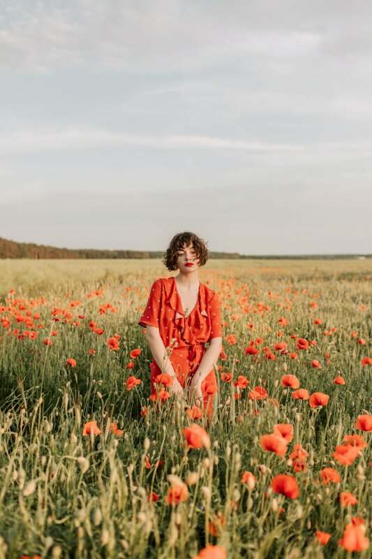 Cách tạo dáng cực chill, cuốn hút của cô gái trẻ trong chiếc đầm dài màu đỏ có mái tóc ngắn hơi xoăn, người hơi nghiêng một bên, 2 tay tạo dáng chạm nhau phía trước tỏa nét đẹp rạng ngời trên cánh đồng hoa dưới nắng vàng nhạt