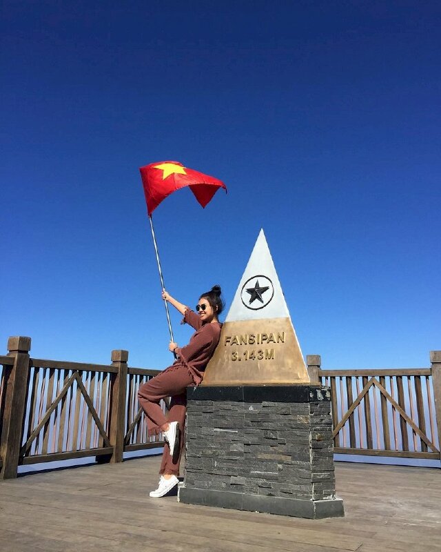 Tạo nét vui tươi với dáng đứng tựa người vào bục Fansipan, 2 tay cầm cờ Tổ Quốc, miệng cười tươi, một chân co tạo dáng