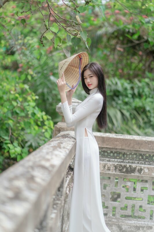 Dáng đứng tự nhiên người hơi tựa vào thành, 2 tay càm nón lá che trước mặt, mặt hướng về ống kính, miệng cười nhẹ