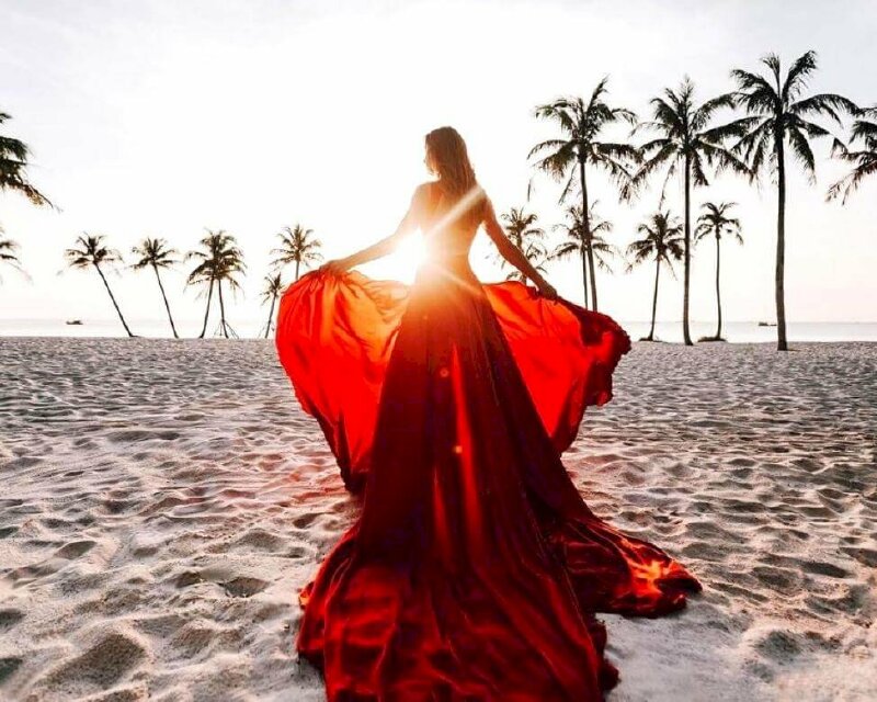 Tạo dáng chụp ảnh với bình minh trên biển cực nghệ thuật của cô gái diện chiếc đầm đỏ dài phết đất, 2 tay nắm váy áo xòe rộng, góc chụp từ sau lưng, mặt hơi nghiêng đón ánh nắng mặt trời, tia nắng xuyên qua tuyệt đẹp