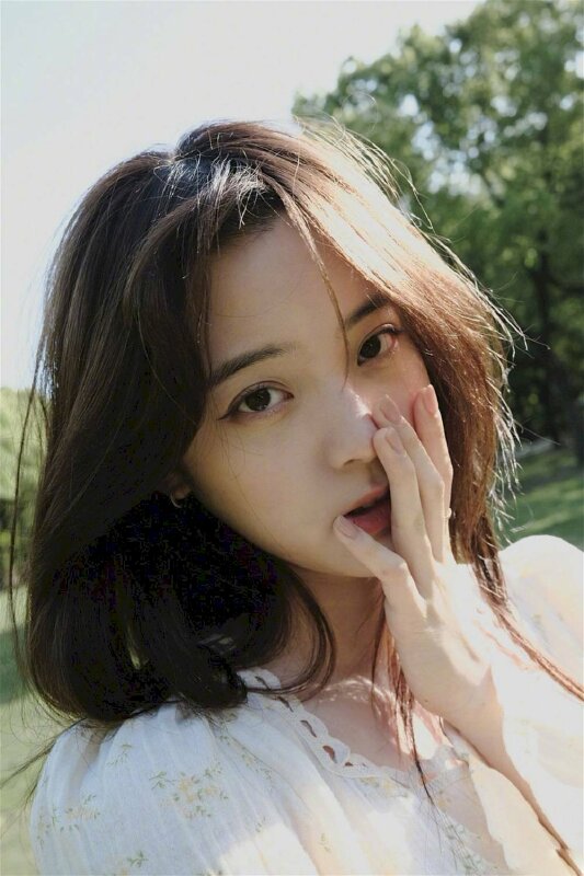 Tạo dáng chụp ảnh cận mặt của cô gái với mái tóc xõa ngang vai, một tay chạm lên má, mắt nhìn ống kính, đầu hơi nghiêng tạo nét hồn nhiên