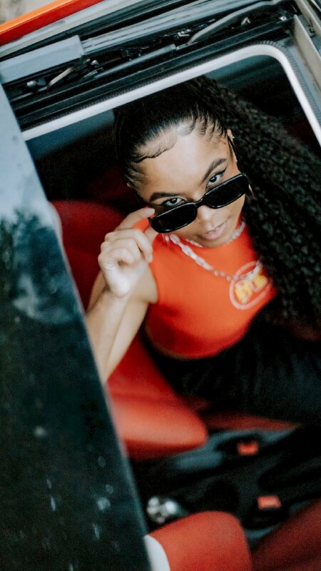 Kiểu dáng ngồi sang trọng của cô gái trong chiếc áo cam cùng chiếc kính đen cực chất ngòi trong xe ô tô với góc chụp từ trên xuống thể hiện chất quý bà vô cùng cá tính