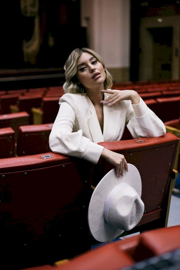 Cách tạo dáng trong rạp chiếu phim khi đèn sáng của cô gái diện chiếc áo trắng ngồi quay ngược về sau ghế, một tay cầm nón, 1 tay chống lên ghế tạo kiểu, đầu hơi nghiêng, mắt nhìn ống kính tạo thần thái khả ái, quyến rũ