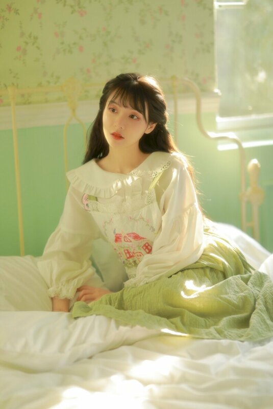 Tạo dáng chụp ảnh trên giường rất dễ thương của cô bé diện đầm trắng, tóc xõa dài, ngồi kê tay lên gường, người hơi nghiêng, dầu ngầng nhìn rất đáng yêu
