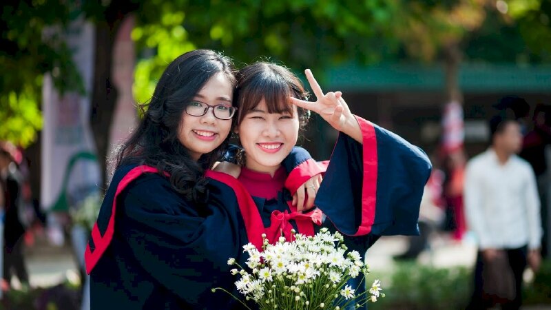 Tạo dáng đẹp trong bộ đồ cử nhân cho 2 bạn sinh viên nữ với kiểu chụp nửa người, một bạn tay tựa lên vai bạn thân, bạn còn lại đưa tay "say hi", miệng cười tươi, mắt nhìn ống kinh