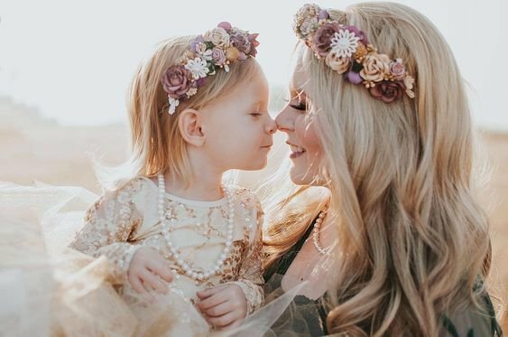 Cách tạo dáng chụp ảnh mẹ và con gái nhỏ đáng yêu khi cả hai mẹ con cùng mặc váy, đội vòng hoa trên đầu, mẹ bồng con gái, đưa con lên mặt con và mặt mẹ quay lại nhìn nhau mũi mẹ chạm lên mũi con, tạo nên cảm giác gần gũi và thân thiết giữa mẹ và con gái.