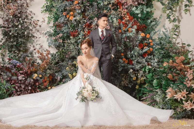 Cách tạo dáng chụp ảnh cưới phim trường đơn giản với bức tường đầy hoa cổ kính, cô dâu ngồi trên ghế, 1 tay đặt lên ngực, 1 tay còn lại cầm bó hoa để trước váy, chú rể đứng phía sau cô dâu 1 tay để vào túi quần. 