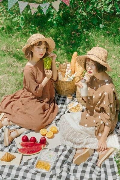 Tạo dáng chụp ảnh đôi bạn thân nữ ngồi trên thảm khi đi picnic với tư thế ngồi 1 bên, góc chụp từ trên xuống, tay 1 bạn cầm trái cây, 1 bạn cầm đồ ăn đưa lên chụp hình. 