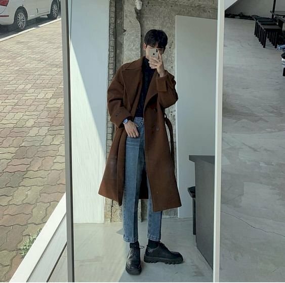 Cách tạo dáng chụp ảnh cho nam gầy khi chụp trước gương với trang phục quần jean, áo giữ nhiệt và áo khoác măng tô dài, hãy đứng thẳng, đầu hơi nghiêng về 1 bên, 1 tay để ở túi quần, 1 tay cầm điện thoại chụp hình, bàn chân để hình chữ V.