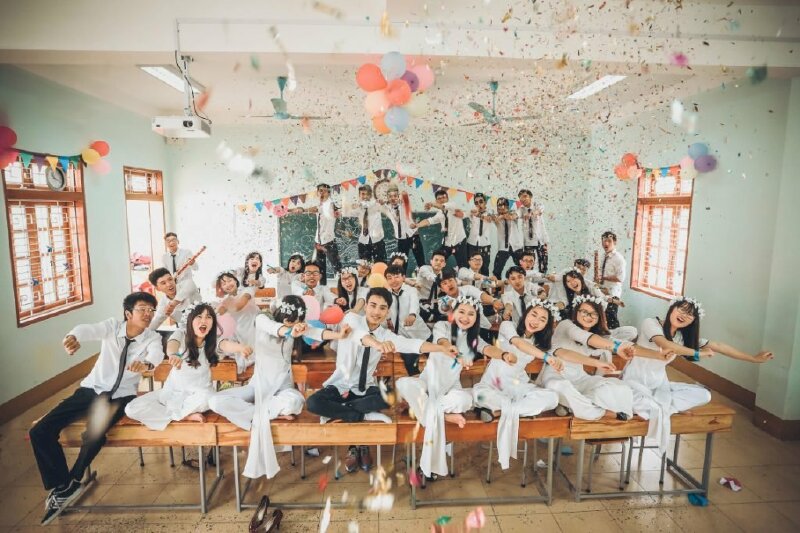 Cách tạo dáng chụp kỷ yếu nhóm lớp dành cho học sinh cấp 3 cuối cấp, tất cả mọi người cùng ngồi lên bàn xếp bằng, đưa tay như đang nhảy vũ điệu cồng chiêng, thể hiện nét đáng yêu, tinh nghịch của học sinh.