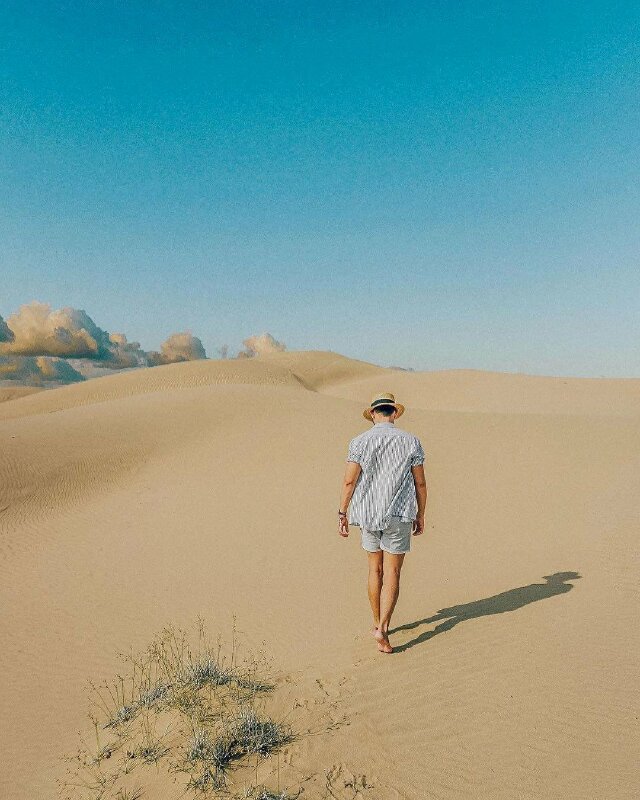 Tạo dáng chụp ảnh trên đồi cát cho nam với dáng quay lưng bước đi tự nhiên trên cát, bước 1 chân trước và 1 chân sau, đầu hơi cúi xuống nhìn đường đi.