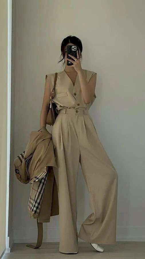 Tạo dáng chụp trước gương với bộ vest màu kem cá tính bằng cách đứng thẳng người chân tạo hình chữ D với 1 chân thẳng, 1 chân hơi cong lên, 1 tay cầm áo khoác và 1 tay cầm điện thoại chụp hình.
