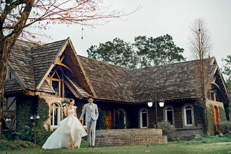 Cách tạo dáng chụp ảnh cưới phim trường với background ngôi nhà gỗ đẹp cổ kính phía sau, cô dâu chú rể đang cầm tay nhau bước đi lãng mạn. 