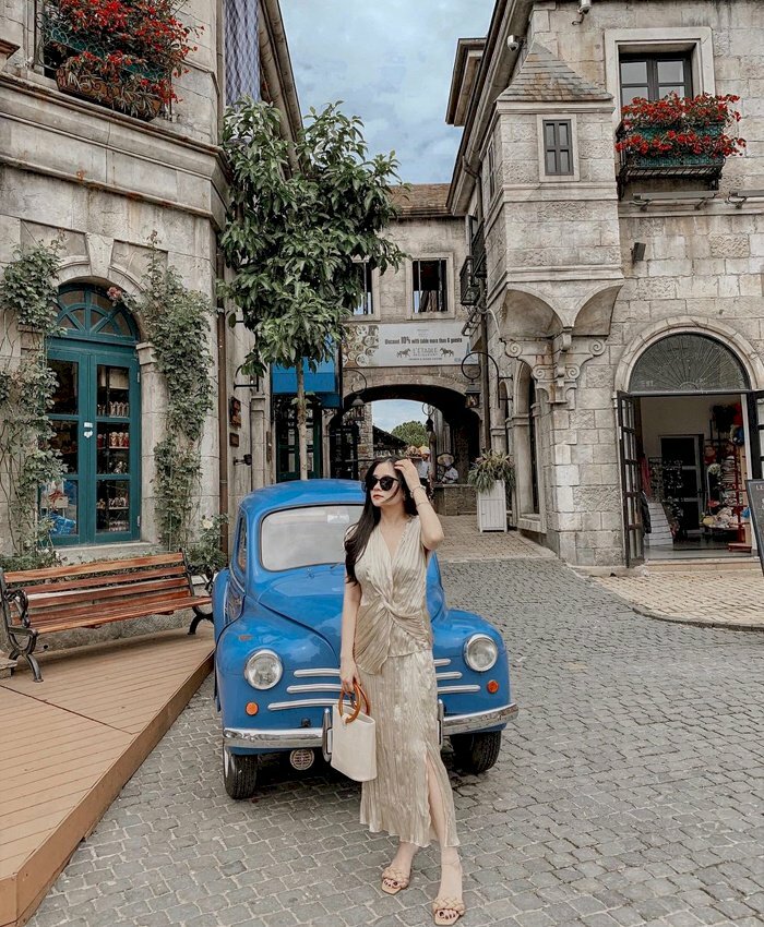 Cách tạo dáng chụp ảnh ở Làng Pháp với chiếc xe ô tô cổ, hãy đứng phía trước chiếc xe, đứng nghiêng người 1 góc 45 độ, đặt 1 chân trước 1 chân sau, 1 tay cầm túi xách, còn 1 tay đưa lên vuốt tóc.