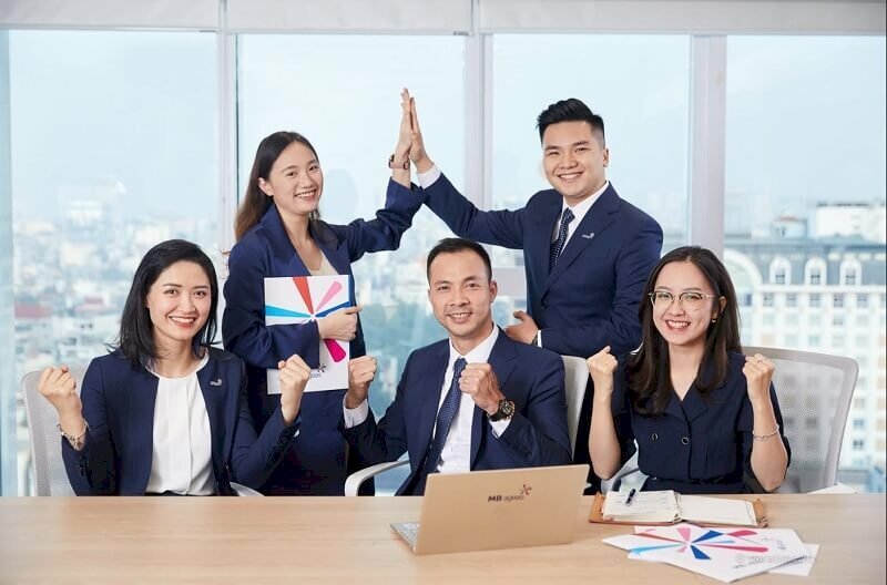 Cách tạo dáng chụp ảnh tập thể công ty cùng nhau cố gắng phấn đấu phát triển, 3 người sẽ ngồi trên bàn làm việc đưa tay nắm chặt hướng lên trời tượng trưng cho sự đoàn kết, sức mạnh, và thách thức và hai người đứng sau đưa tay tạo hình tháp. 