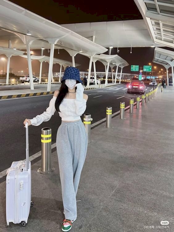 Cách tạo dáng chụp ảnh ở sân bay cho nữ với dáng đứng chéo chân, một tay cầm vali và một tay đưa lên gần chạm nón.